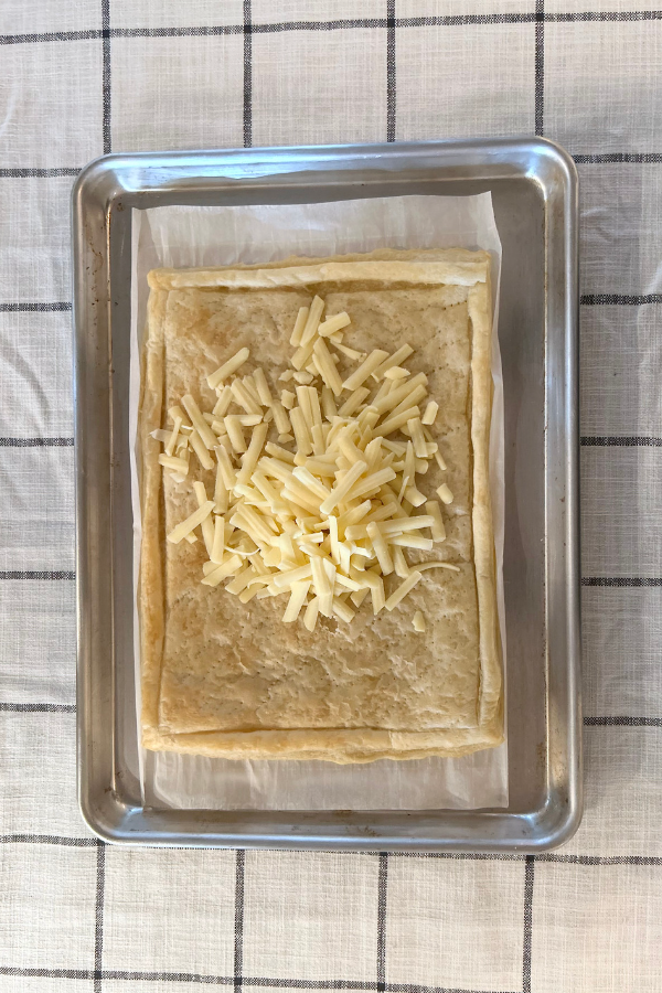 A par-baked pastry shell with a pile of cheese in the middle, placed inside of a rimmed baking sheet on a black and white table cloth.