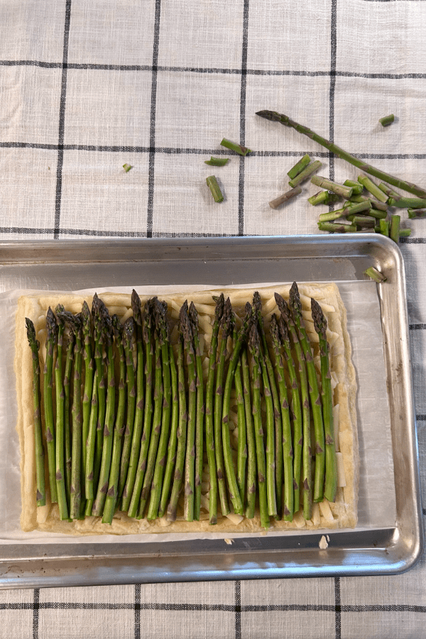 A par-baked pastry shell, covered in cheese and asparagus, placed inside of a rimmed baking sheet on a black and white table cloth.