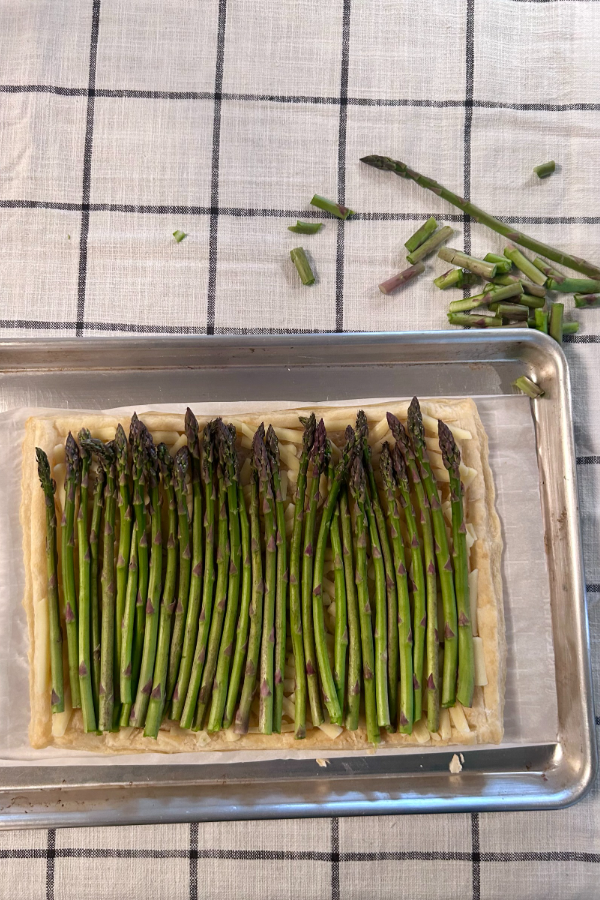 A par-baked pastry shell, covered in cheese and asparagus, placed inside of a rimmed baking sheet on a black and white table cloth.