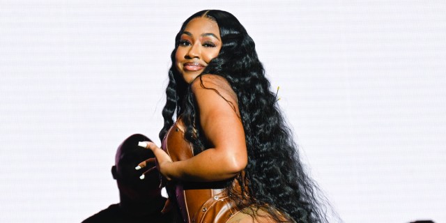 Yung Maimi, who is bisexual and friends with Megan Thee Stallion, performs in a brown leather bodysuit onstage, she is smiling.