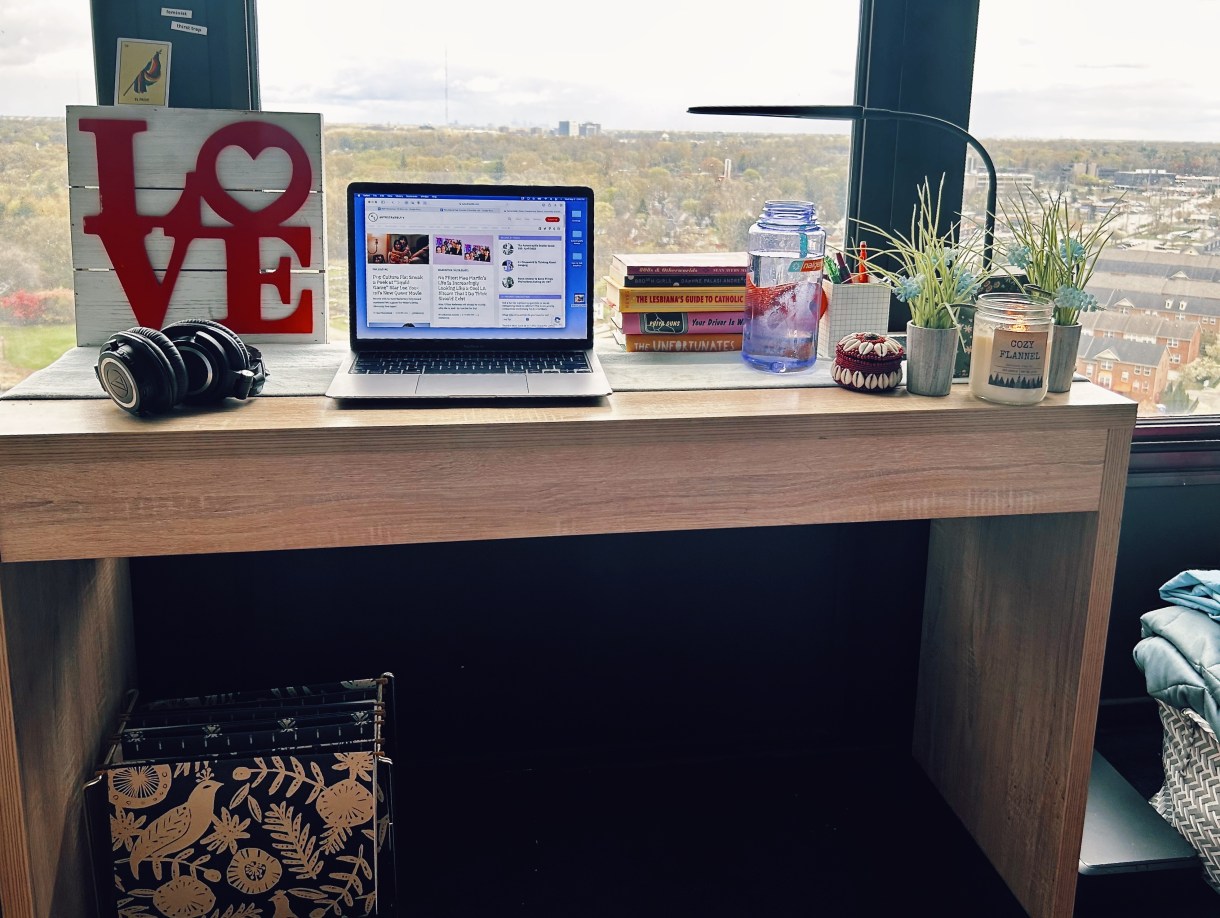 A photo of Carmen's desk against a large bedroom window that overlooks the city below. There are books stacked, plants, a laptop, and some other items, but mostly this is a minimalist desk. Very tidy.