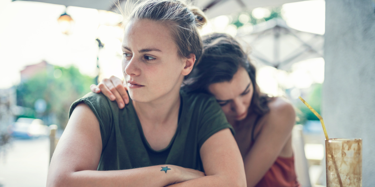 a queer white woman looking pensively into the distance. she has long blonde hair with a grown-out undercut on the sides. a brunette woman with a widow's peak is hugging her from behind.