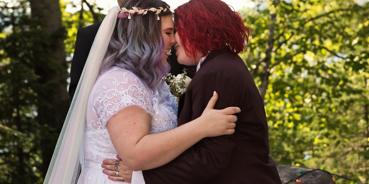 two white lesbian brides — one with blueish dyed hair and one with red hair — embrace during a gay wedding ceremony