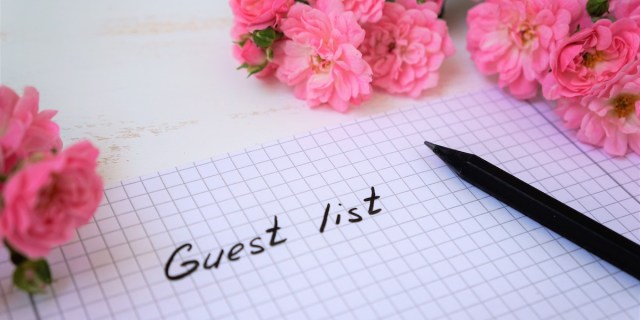 a piece of graph paper that says "guest list" lies on a table with a black pen surrounded by little pink roses