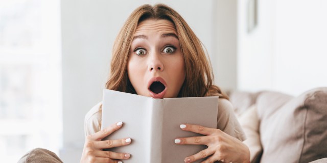 a woman is sitting on the couch reading a book, and she pops her head above the pages to make a truly shocked face: eyes wide, eyebrows raised, mouth open! she's surprised!