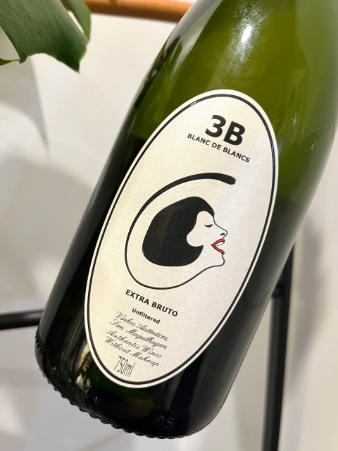 The bottom half of a green bottle of wine. The label has a woman's face in the center. Above the face is text that says "3B BLANC DE BLANCS" and below the face is more text that says "EXTRA BRUTO".