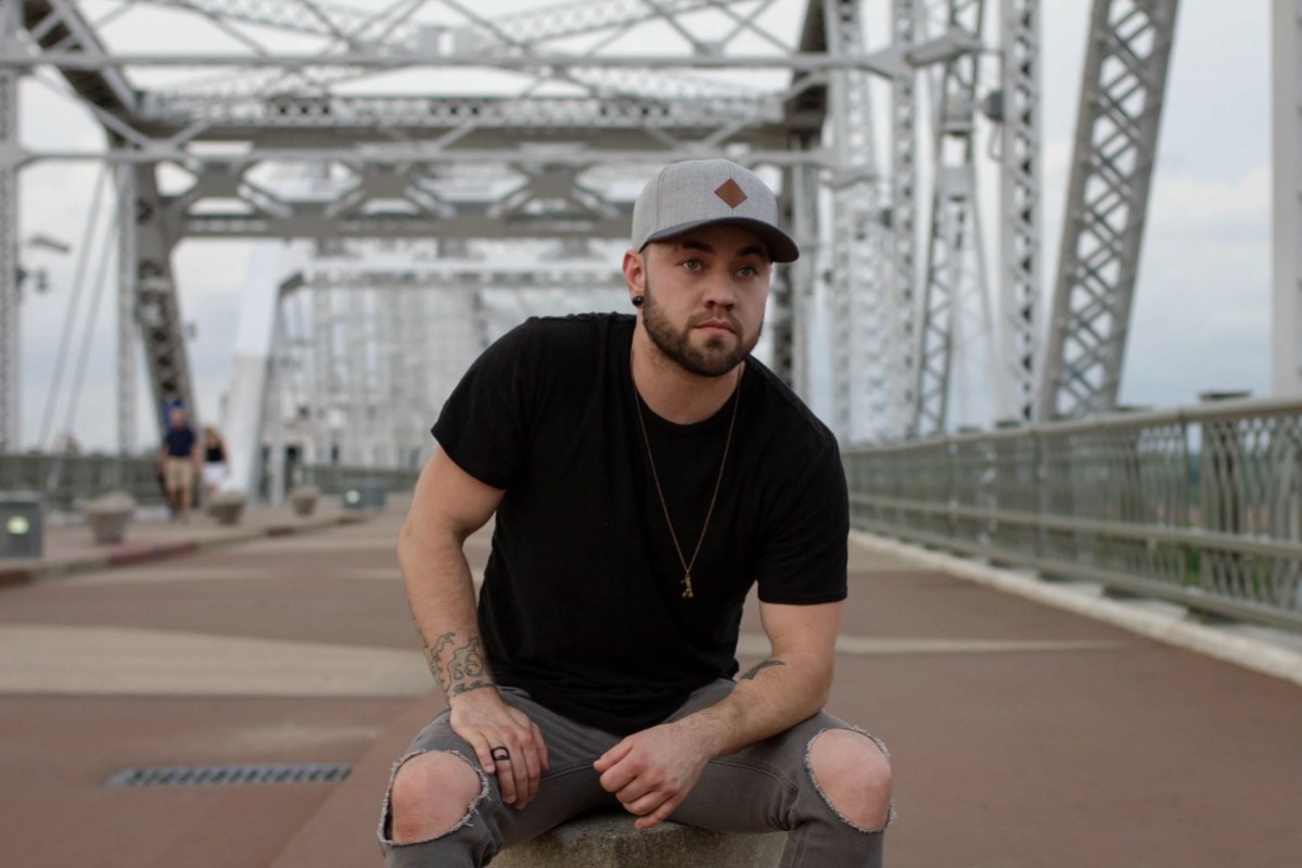 Brody Ray, a transmasc country singer, wears a black tee and flat brimmed hat while crouching on a bridge