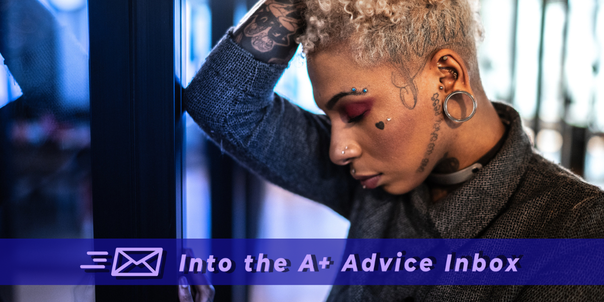 A woman of color in a blazer in a workplace looks pensive. She has bleach blonde curly hair and multiple tattoos and is on the masc end of gender presentation