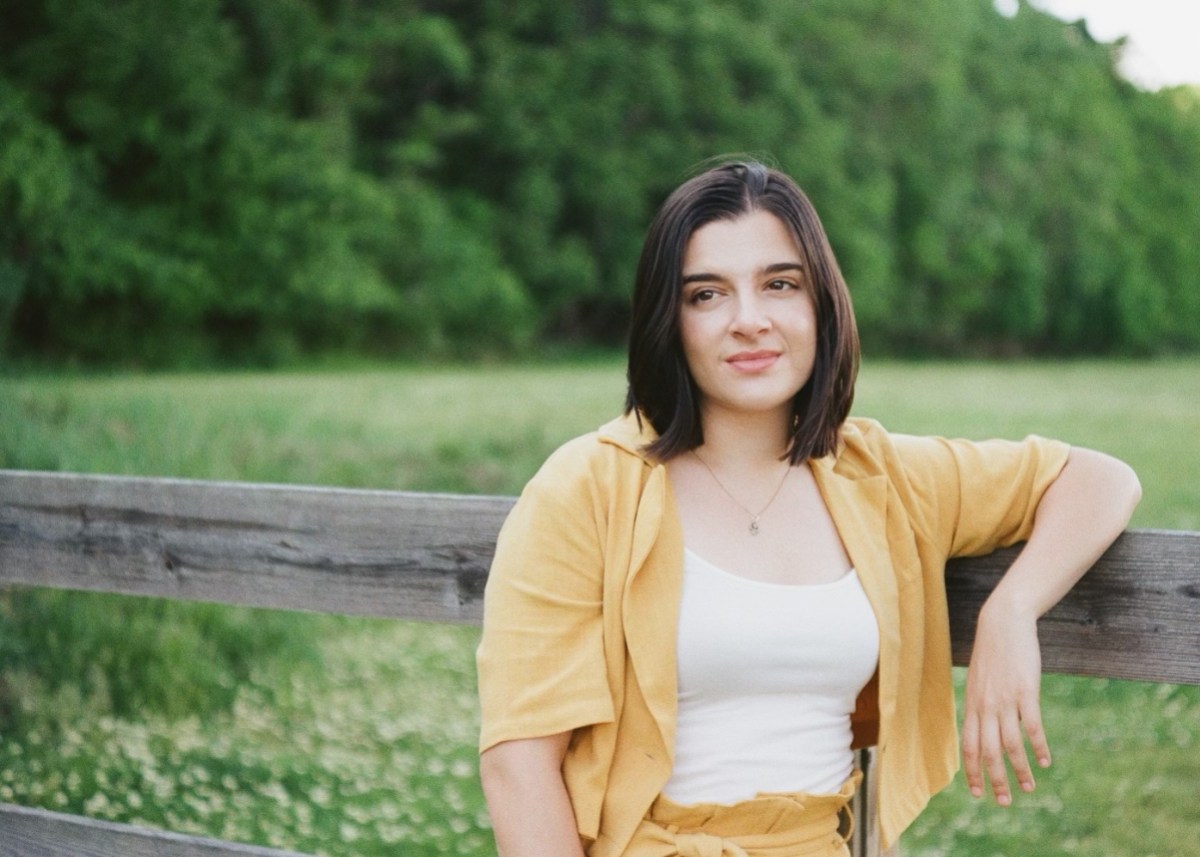 Madeline Kelson the country singer in an orange short sleeve set leans against a fence in a field