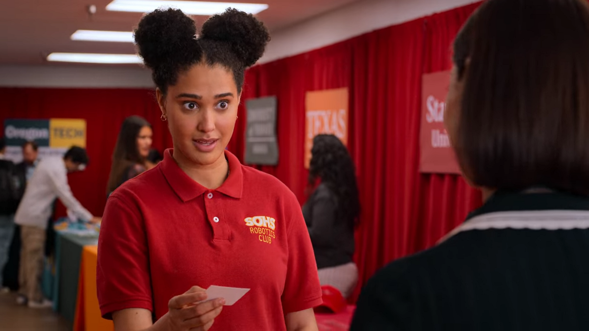 Fabiola is holding a business card and smiling. She's wearing a red polo shirt for the Robotics club and her hair is in buns. We see the back of the Princeton rep, who handed Fabiola her card.