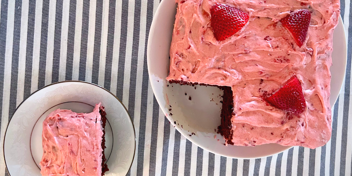 A close up of a chocolate cake with strawberry frosting and fresh strawberry decorations, on a stripped table cloth.