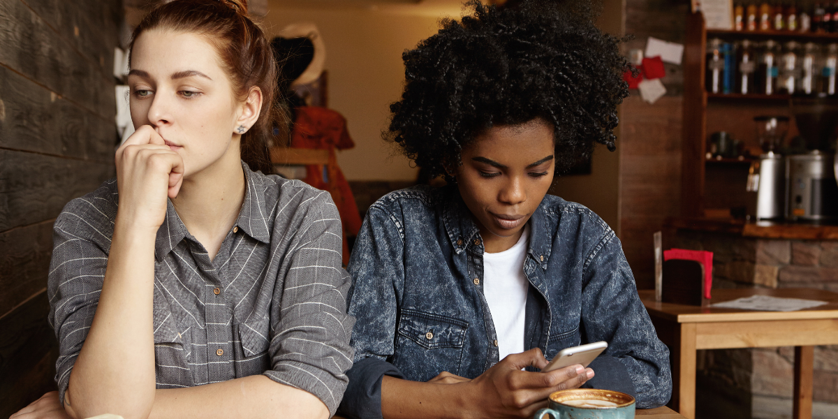 A white woman with long blonde hair in a bun wears a grey flannel with white cross hatching. She sits in a cafe with her chin in her hand, looking away from the woman sitting next to her. The woman beside her is a Black woman with curly hair. She wears a white shirt with an open denim collared shirt over it. She looks down at her phone. A latte is in blue mug in front of her.