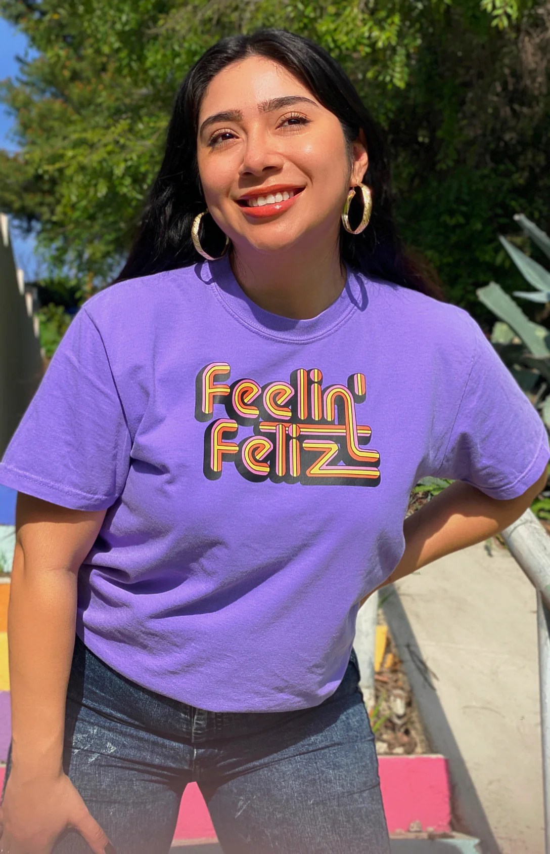 model in a rich purple tee with "Feelin Feliz" in retro font across the chest