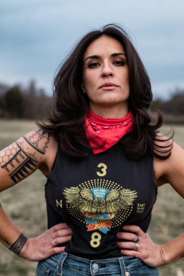 Mercy Bell a queer country singer wears a red bandana and black tank and has arm tattoos and is standing in a field with her hands on her hips