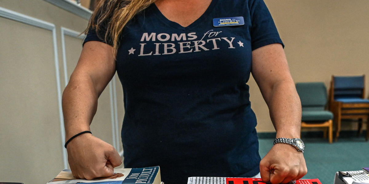 a woman wearing a MOMS FOR LIBERTY t-shirt stands over books