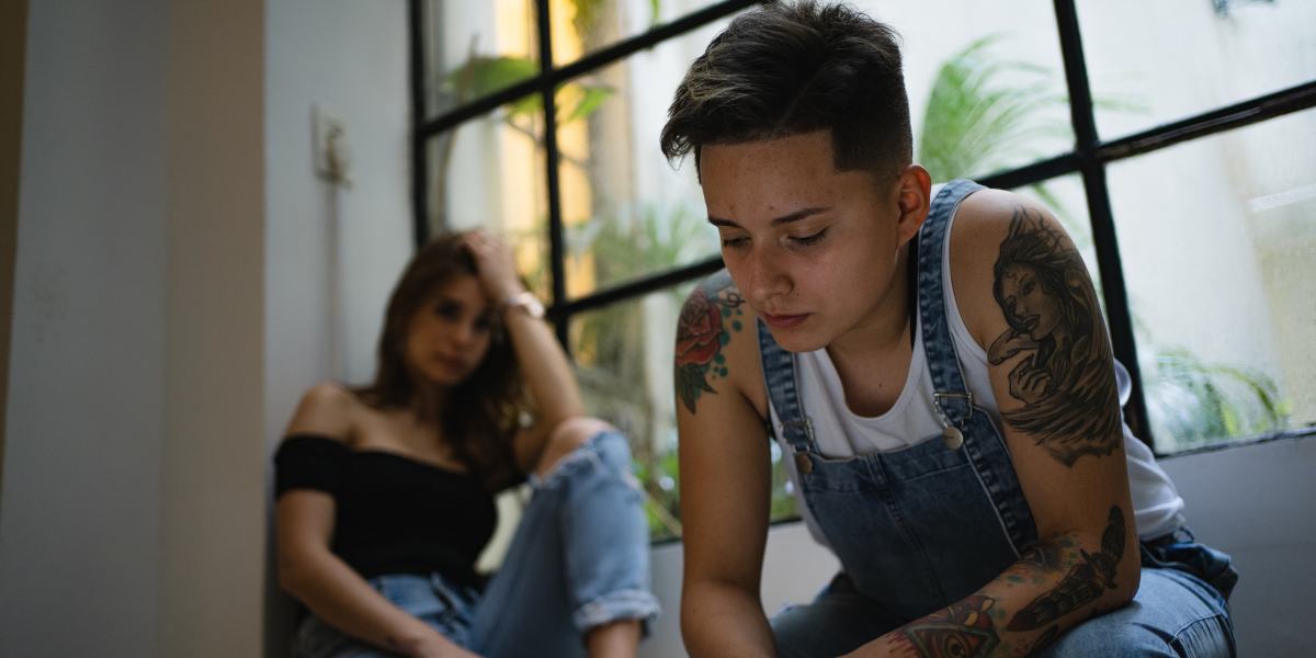 an androgynous person in overalls and tattoos looks sad, sitting next to a femme person with a black top and ripped jeans