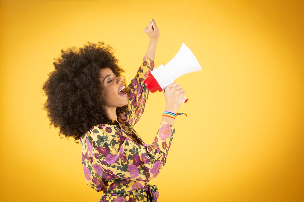 a stock photo of a woman holding a megaphone, rainbow bracelet around her wrist, shouting cheerfully and raising her fist