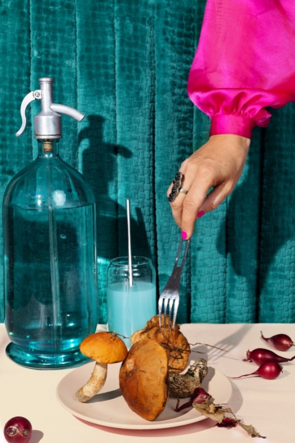 a hand, arm above ensconsed in a bright fuschia sleeve, descends with a fork that the model is sticking into a plate of wild mushrooms