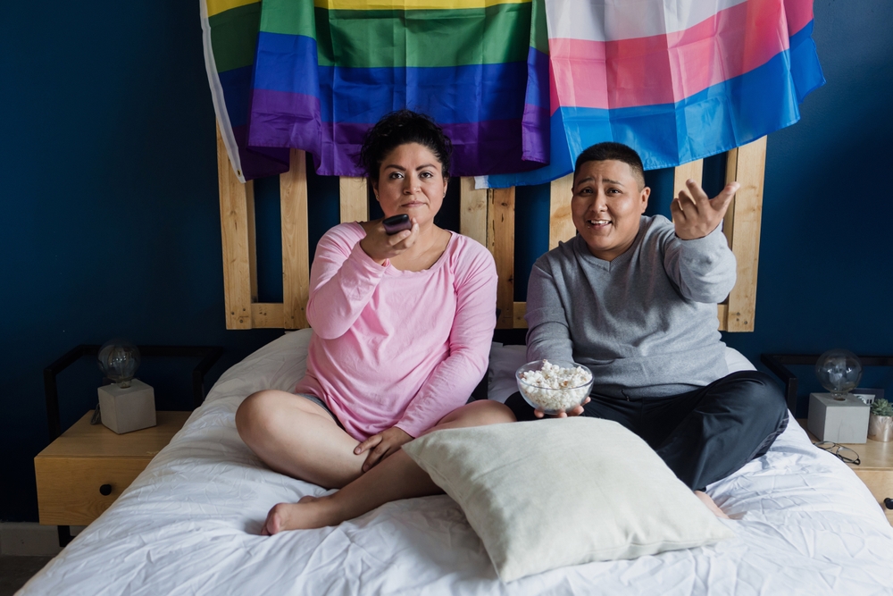 a couple, a woman and a trans man, sit in in bed, pride flags behind them, watching a movie and holding popcorn