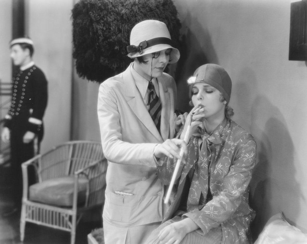a vintage photo of two women, the one in a suit, obviously a capricorn, the most dapper of the signs in this gay pride horoscope, lighting the cigarette of a another woman in 1920s style clothing