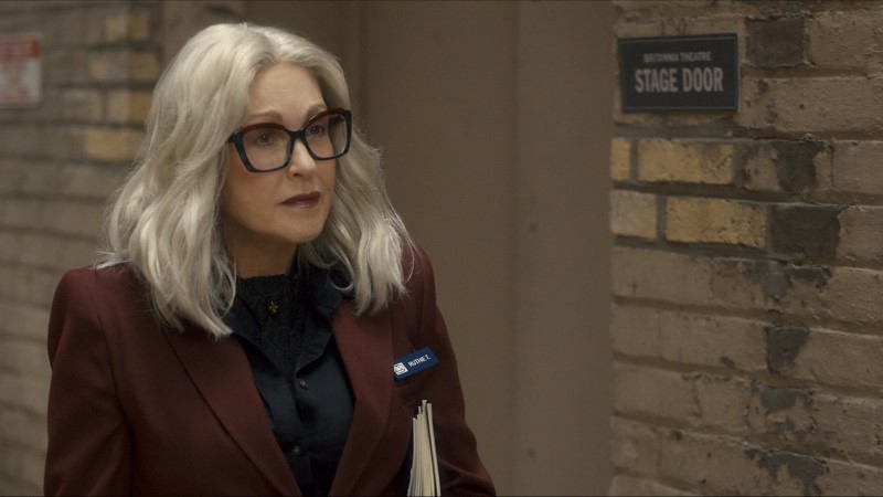 Cyndi Laupre has on a silver shoulder length wig and thick black glasses, plus a burgundy blazer, as Ruthie. She is in a back back alley.