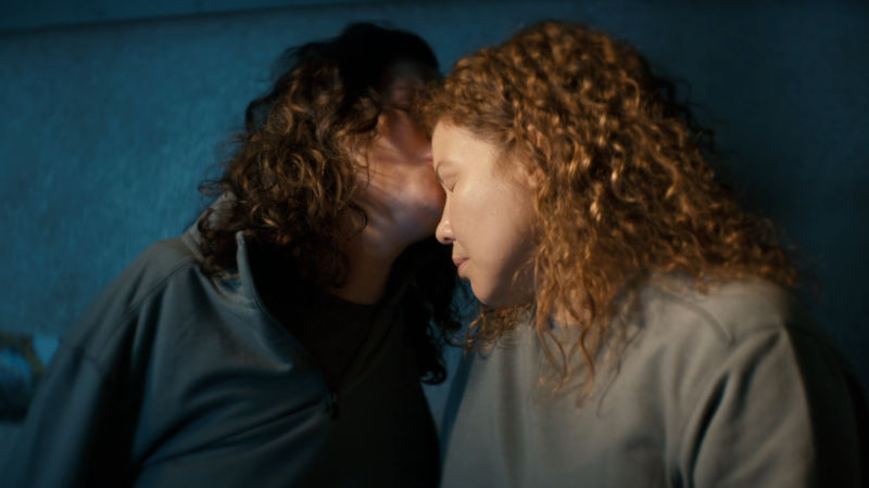 Two women with curly hair, a brunette white woman and a Latina, tenderly hold each other and share a forehead kiss against a dark wall.