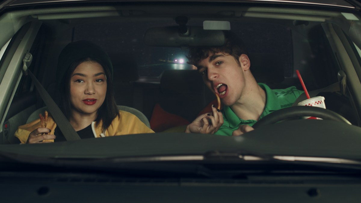 An Asian teen girl and a white teenage boy share a French fry in a car at night. 