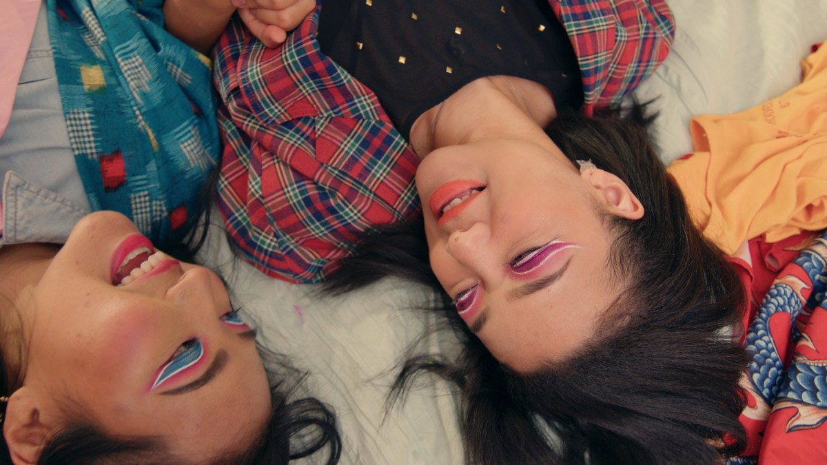 Two Asian teens with detailed, bright eye make up, lay together on a bed.