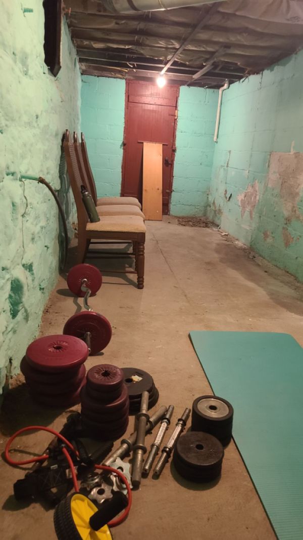 a photo of nico's workout space which is a bunch of barbell and dumbell stuff and plates on a dirty basement floor with crumbling paint