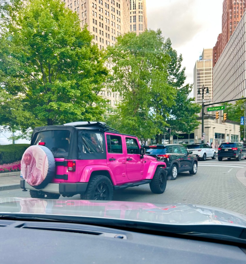 A hot pink jeep is driving around downtown Detroit, on the back wheel is a pink wheel cover with a rapper on it.
