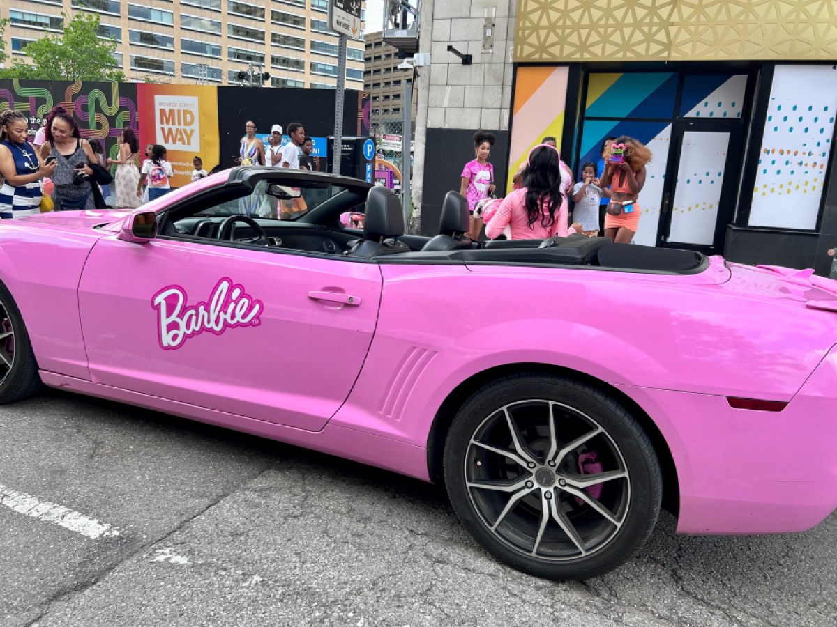 A pink corvette says "barbie" on the side in downtown Detroit, people are taking pictures beside it.