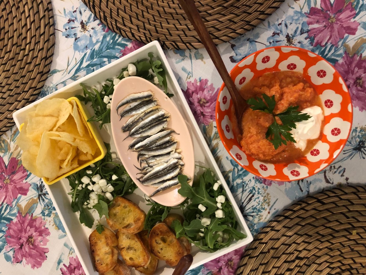 a bowl of gazpacho and a snack plate with arugula, goat cheese, potato chips, and crostini on it