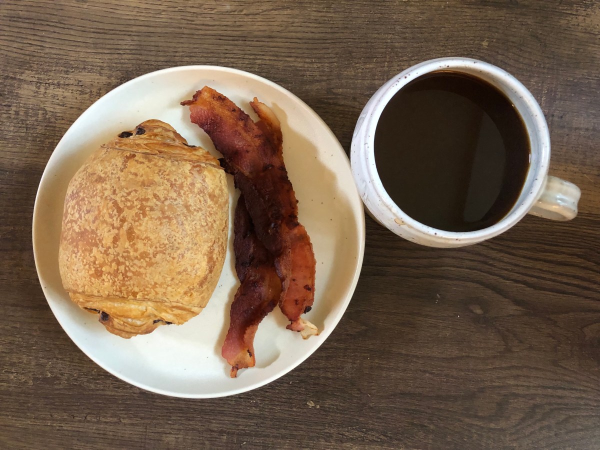 a plate with a chocolate croissant and bacon on it, next to a mug of coffee
