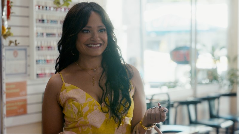 Judy Reyes has wavy hair past her shoulders as Marcie, and is smiling in a yellow spaghetti-strapped sundress. 