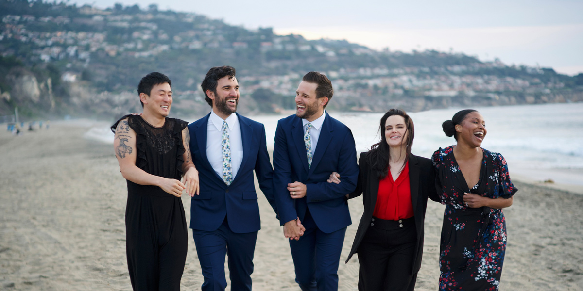 A collection of queer people in formal wear walk hand in hand on a beach during a cloudy day, they are of different genders and races.