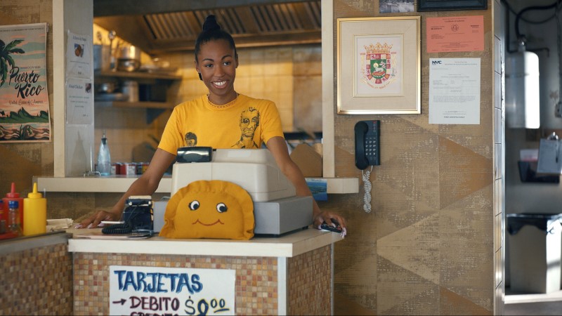 Kita Updike as Nellie is a Black trans woman with a high ponytail in a braid, she's working the register at an empanada shop in a yellow t-shirt.