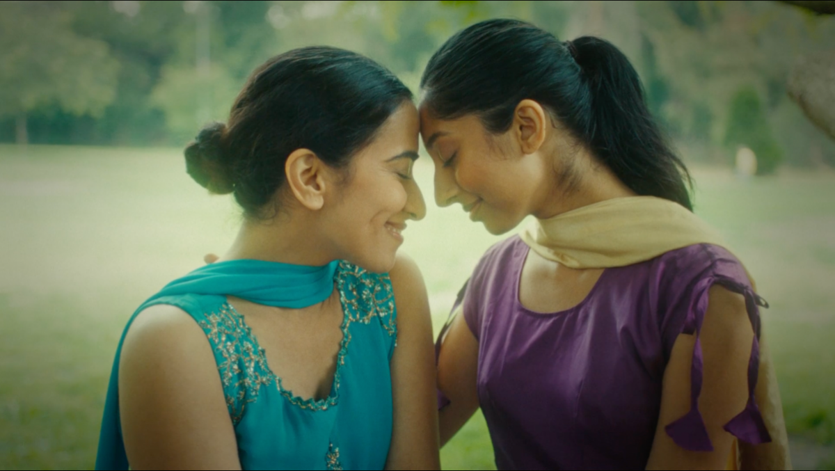 Two young South Asian women touch foreheads tenderly in a field.