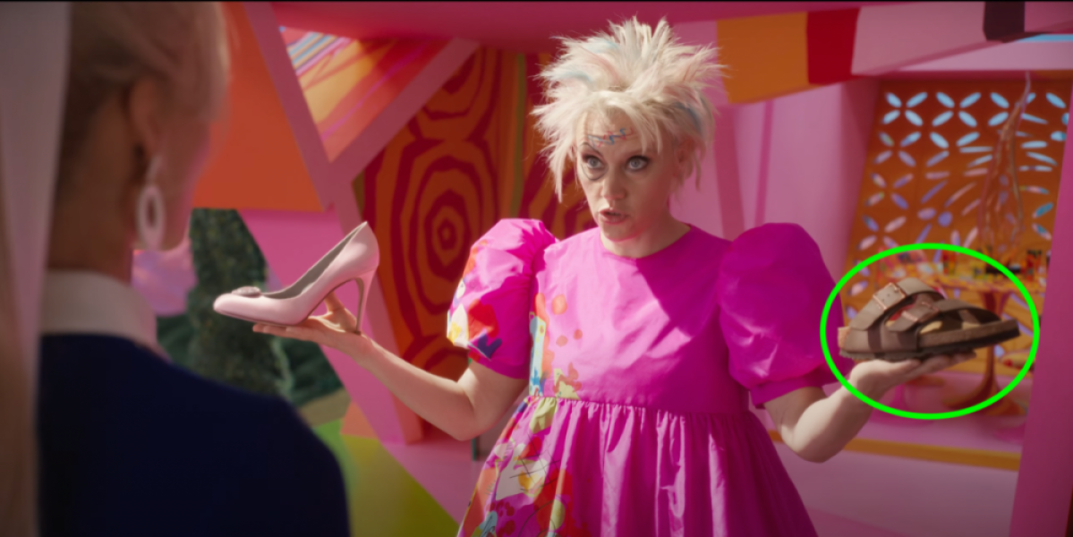 Kate McKinnon in the Barbie movie holding up a pink heel and a birkenstock, imploring margot robbie to choose