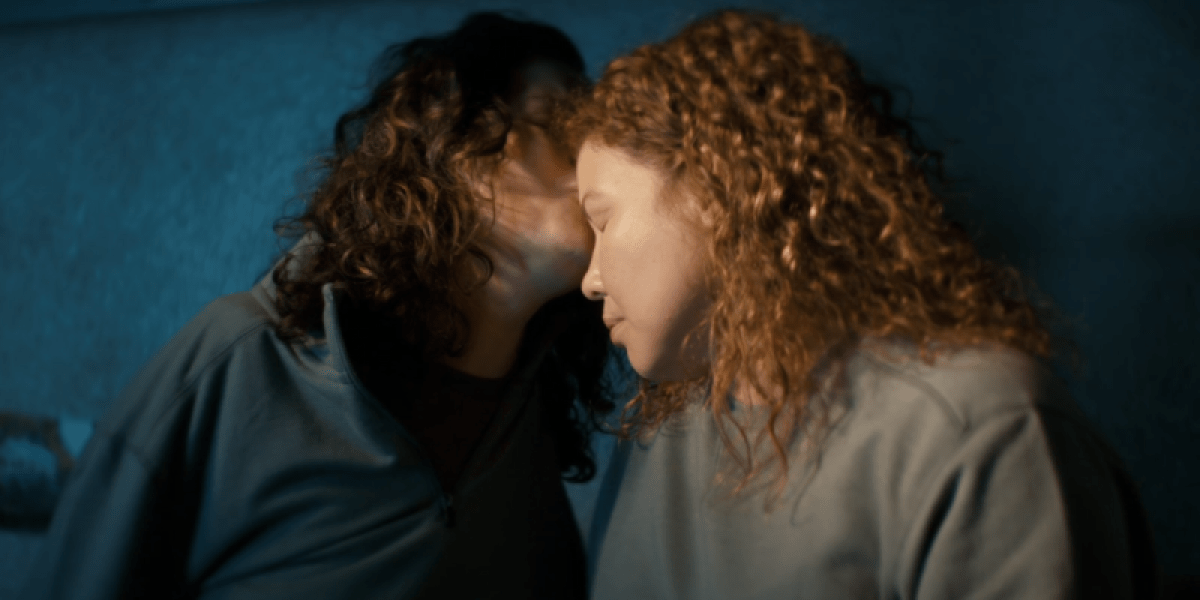 Two women with curly hair, a brunette white woman and a Latina, tenderly hold each other and share a forehead kiss against a dark wall.