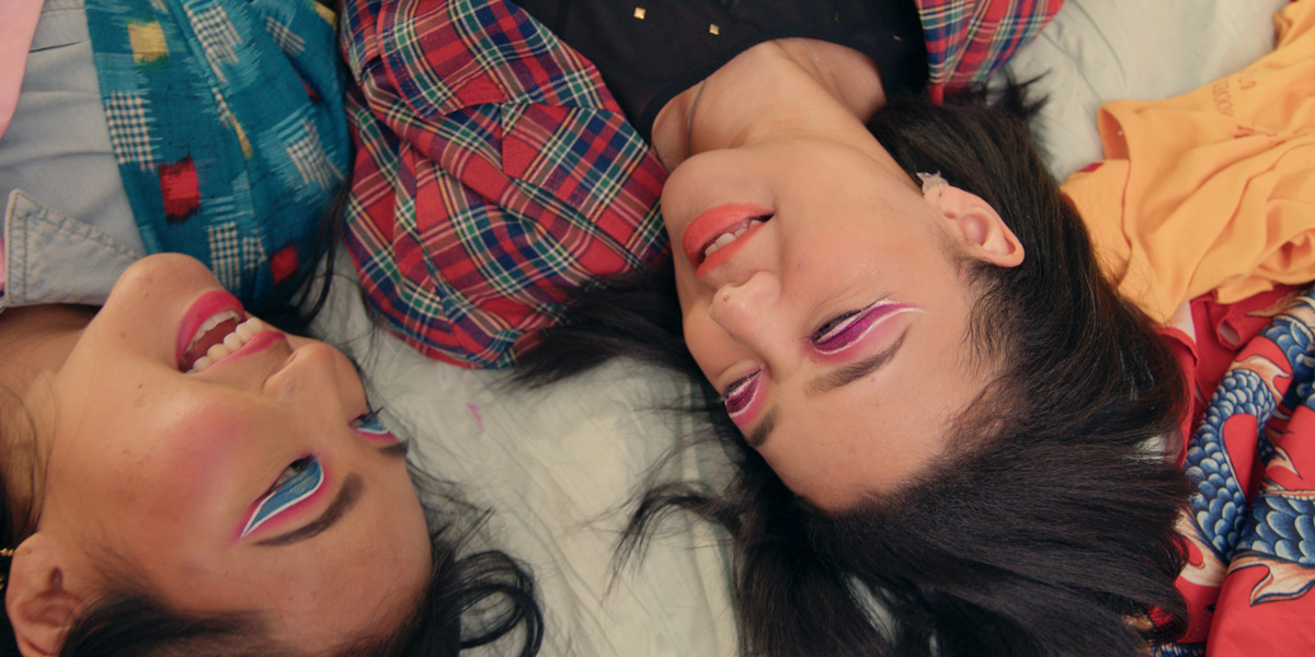 Two Asian teens with detailed, bright eye make up, lay together on a bed.
