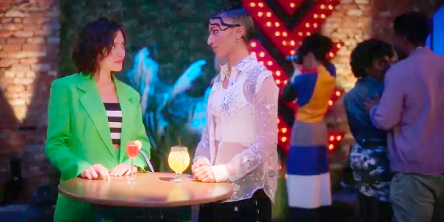 While at the gallery, Drea and Zara enjoy drinks, while standing, at a bistro table. Drea is on the left, wearing a green blazer with matching pants and a black and white top. Zaara is on the right wearing a white tank covered by see-through silk organza shirt.