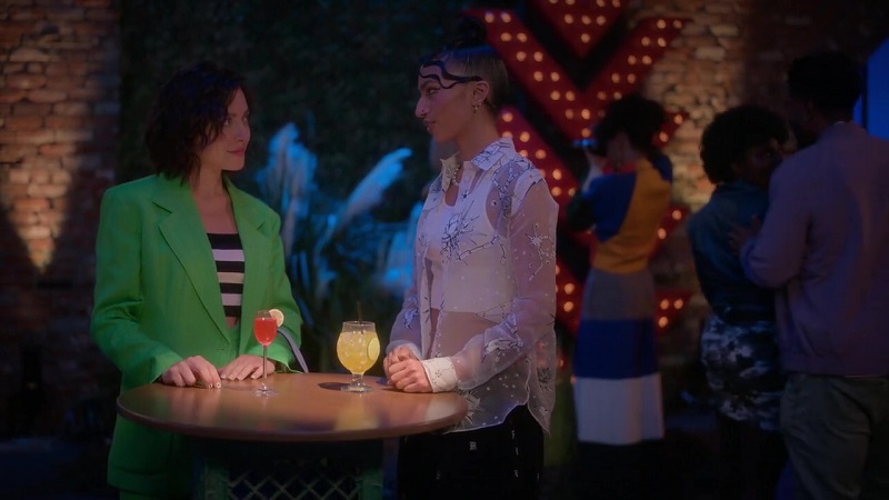 While at the gallery, Drea and Zara enjoy drinks, while standing, at a bistro table. Drea is on the left, wearing a green blazer with matching pants and a black and white top. Zaara is on the right wearing a white tank covered by see-through silk organza shirt. 