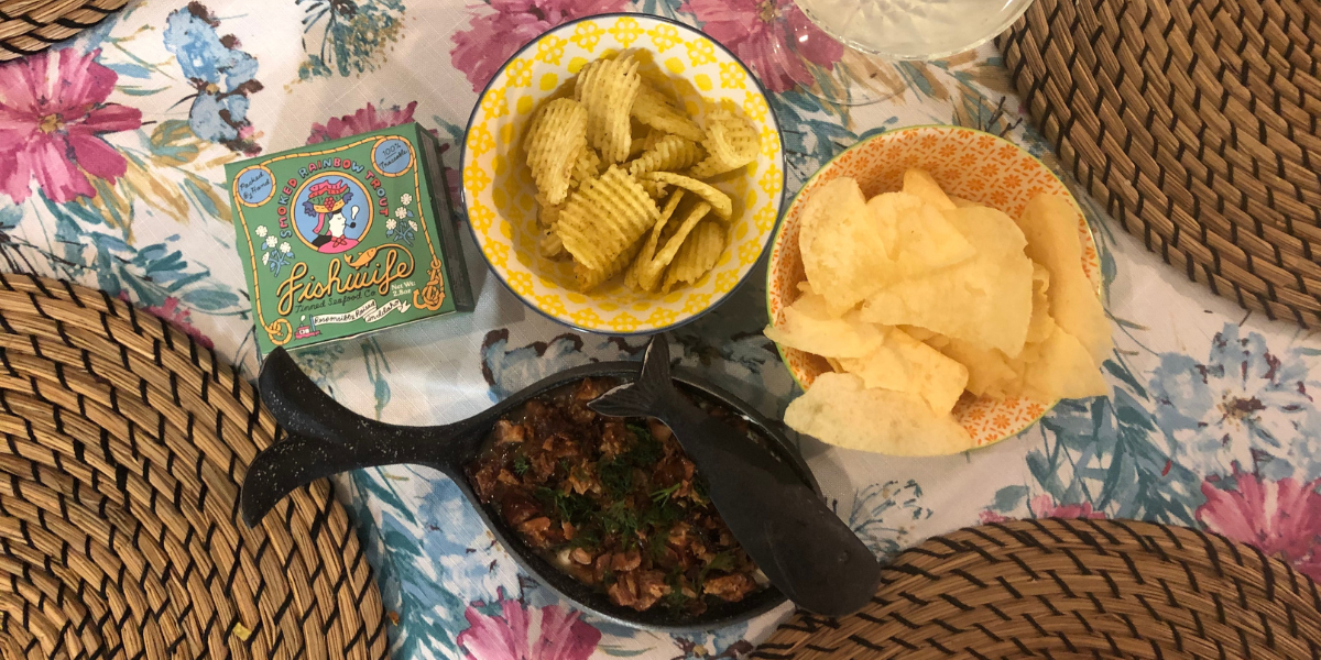 two small bowls of potato chips, a Fishwife rainbow trout tin, and a dish shaped like a fish with trout dip in it