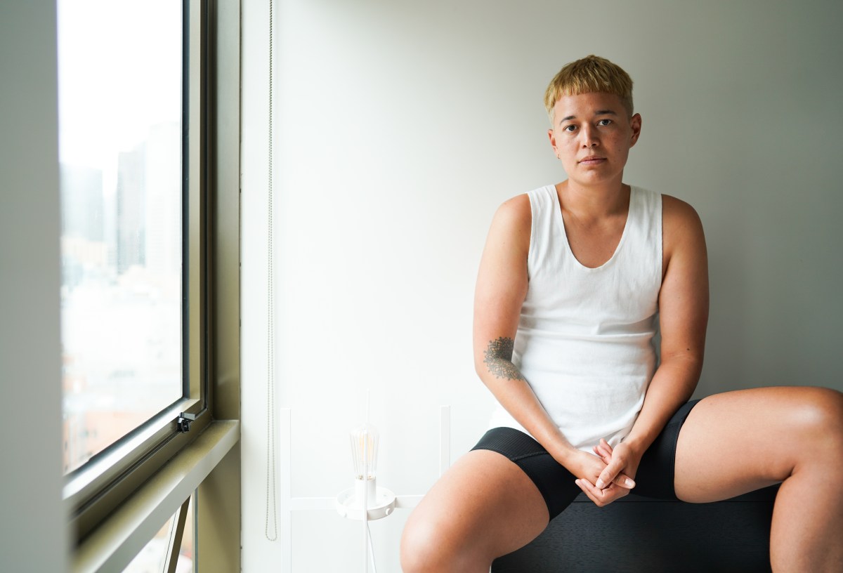 a butch with short bleached blonde hair sits in a white tank looking at the camera