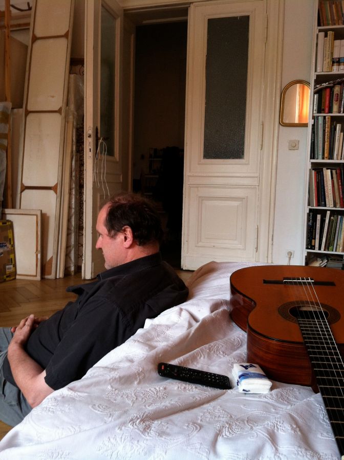 a man sits with his back against a bed and a guitar on it