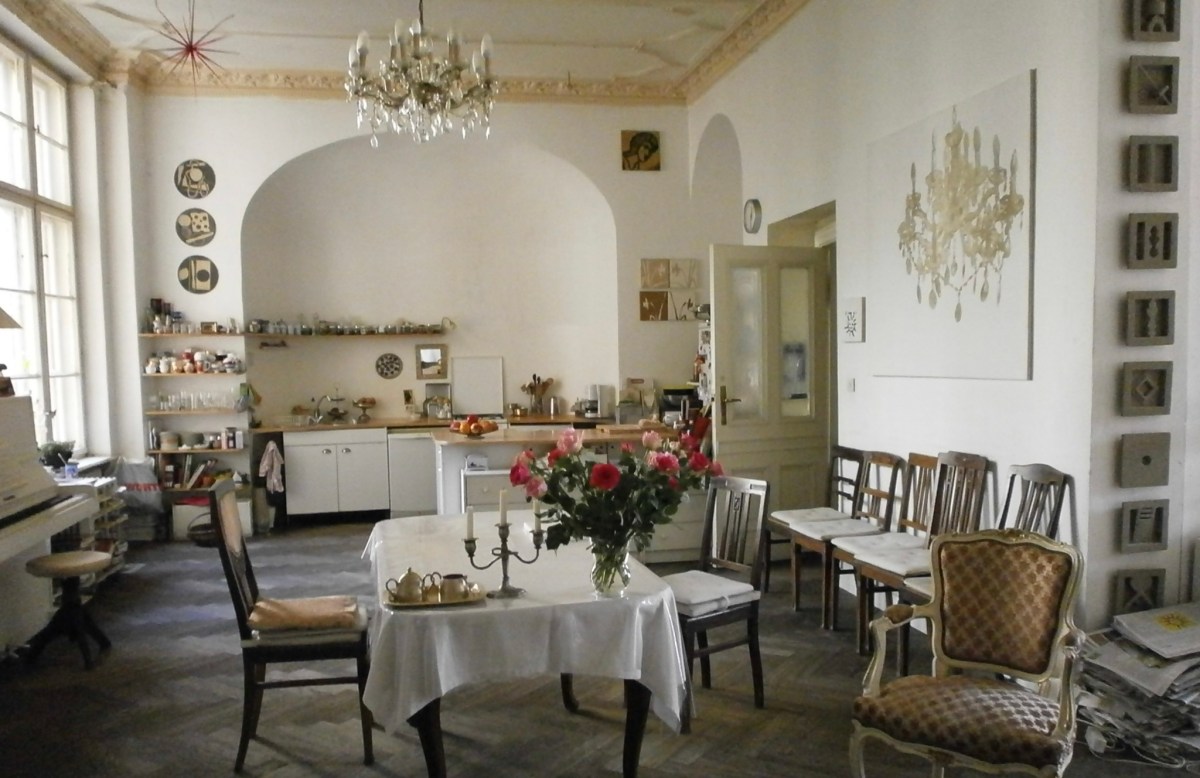 the author's host home in Berlin. a kitchen eating area with a chandelier, a table with a white tablecloth and flowers on it, rustic looking chairs, white walls.