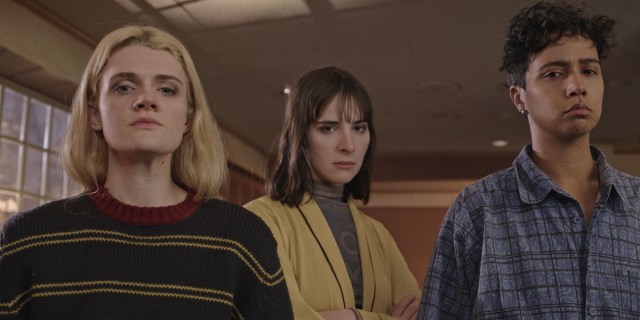 Gayle Rankin, Hari Nef, and Rad Pereira stare toward the camera with concern. Rankin wears a striped sweater, Nef a yellow blazer, and Pereira a blue patterned button down.