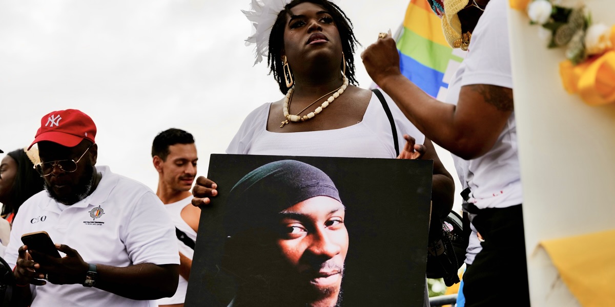 Queen Jen, dressed in all white, stands while holding a sign honoring O’Shae Sibley at his memorial on August 04, 2023 in New York City. The memorial was held at the gas station where he was murdered last weekend while dancing with friends.