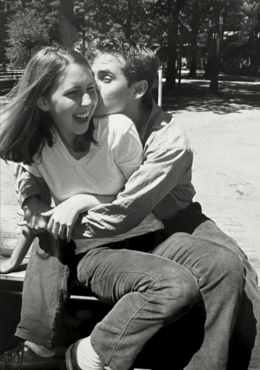 a black and white photo of Riese laughing while sitting wrapped up in the arms o fher gay boyfriend. they are outside.