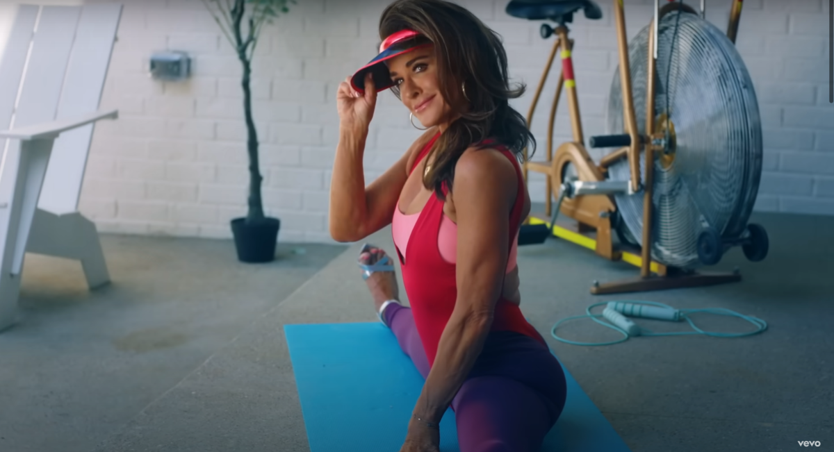 Kyle Richards tipping a visor while in the splits and wearing 80s garb in the Fall in Love With Me music video